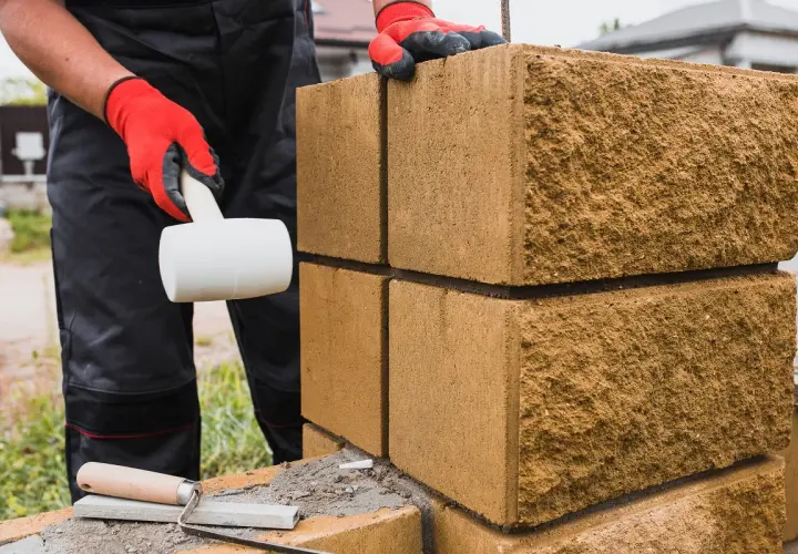 Budowa płotu z bloczków betonowych: ręka w czerwonej rękawicy z młotkiem gumowym i kielnia.