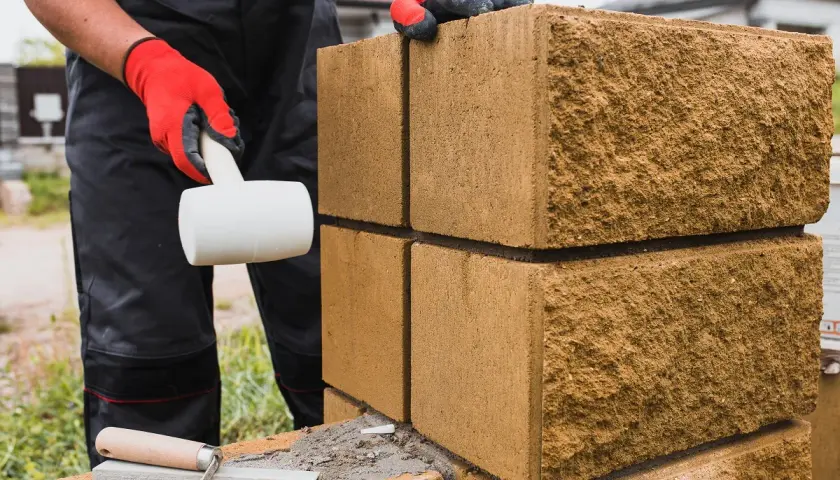 Budowa płotu z bloczków betonowych: ręka w czerwonej rękawicy z młotkiem gumowym i kielnia.
