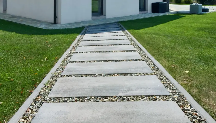Ścieżka z prostokątnych płyt betonowych i kamieni ozdobnych obok trawnika i białego domu.
