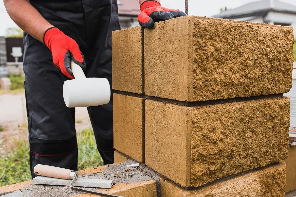 Budowa płotu z bloczków betonowych: ręka w czerwonej rękawicy z młotkiem gumowym i kielnia.
